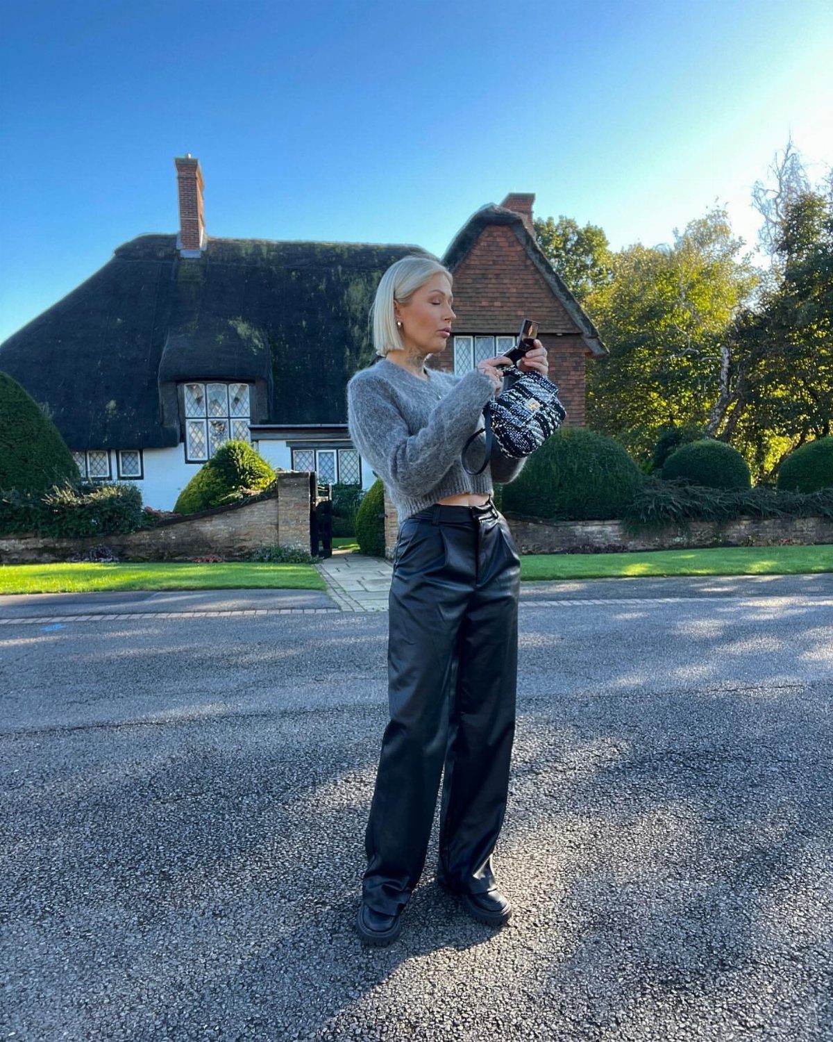 Blonde woman wearing a grey knit sweater and black leather trousers standing outdoors.