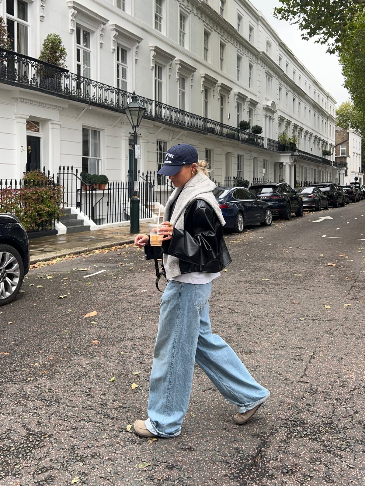 A woman in a leather jacket and wide-leg jeans walks along a residential city street.