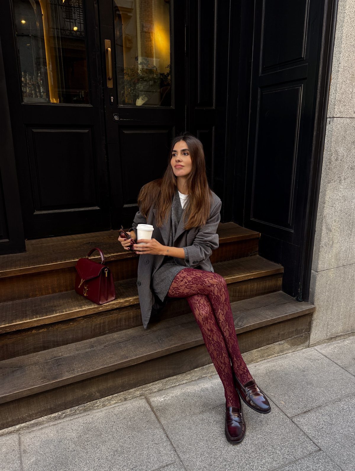 A woman sitting on steps wearing a grey blazer and patterned burgundy tights with loafers.