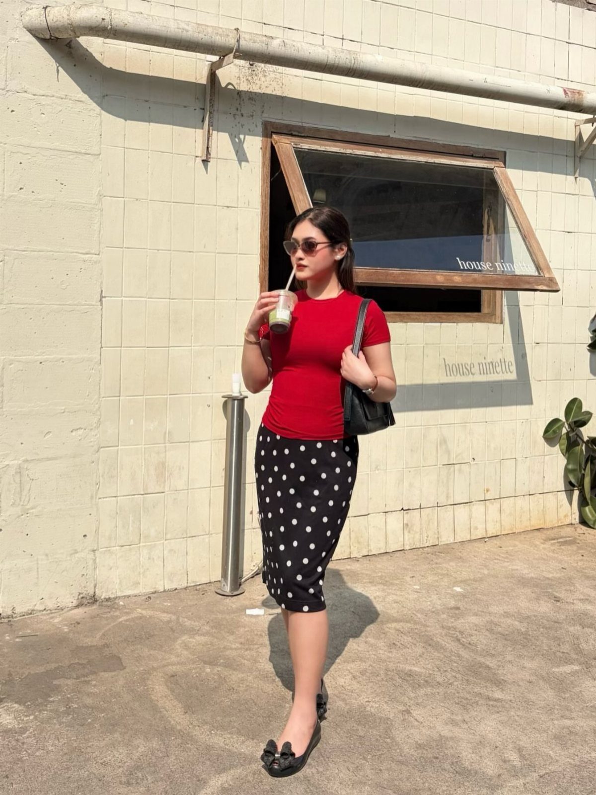 Woman wearing a red top and black polka-dot skirt standing against a white wall.