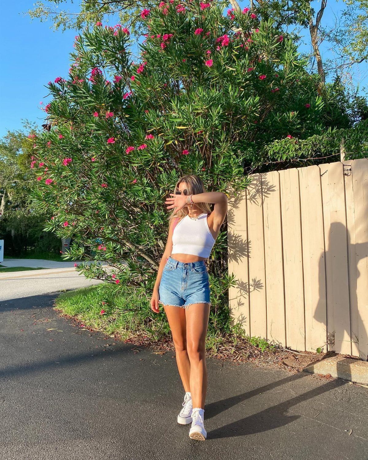 A woman wearing a white crop top and denim shorts standing by a flowering bush.