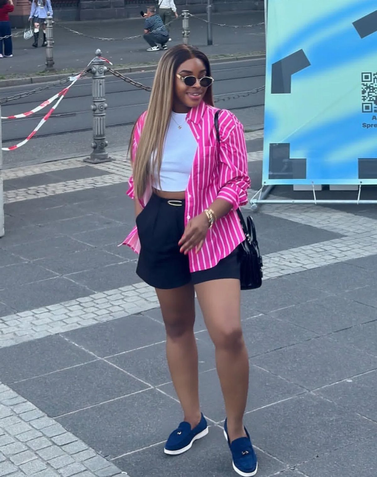 Woman wearing a pink striped button-down shirt, white top, black shorts, and navy loafers.