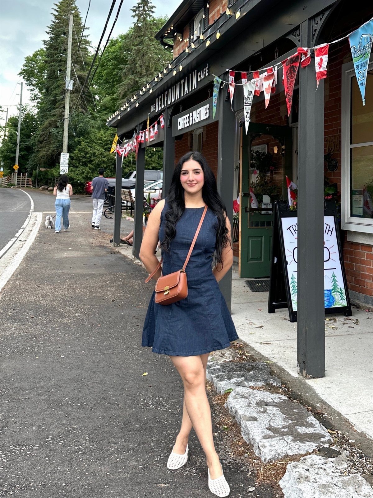 A woman in a dark denim dress posing on a sidewalk in front of a cafe.