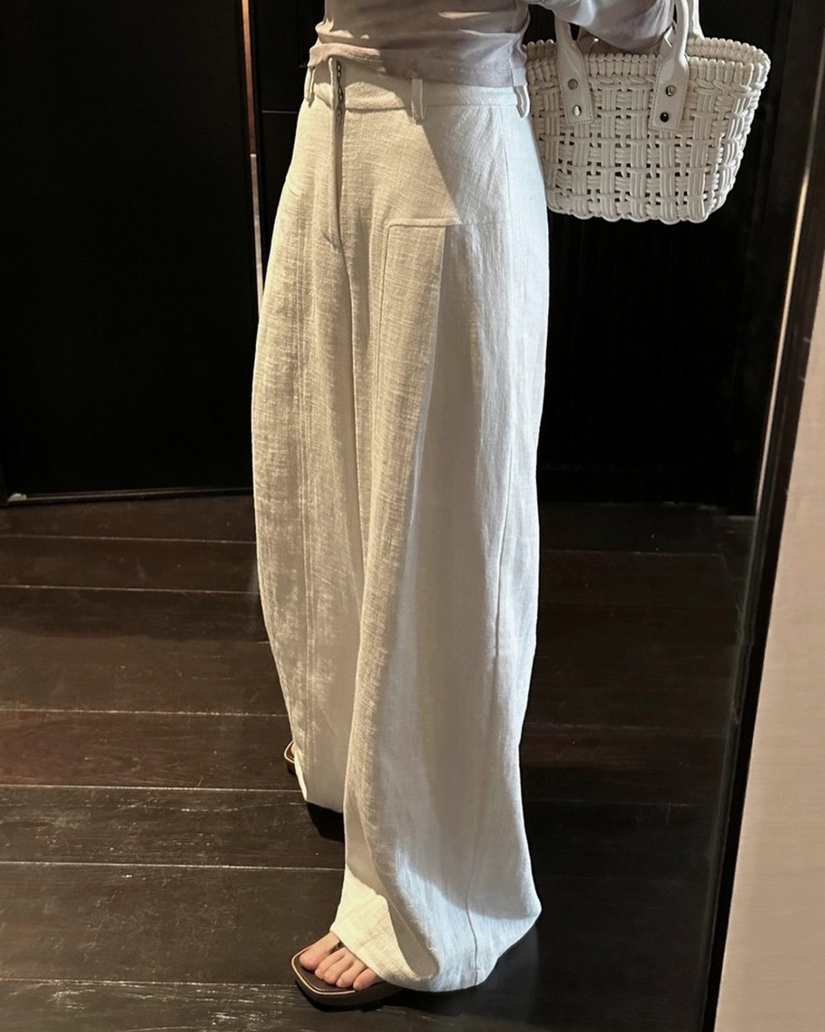 A person wearing flowing white wide-leg linen pants and brown sandals holding a woven bag.