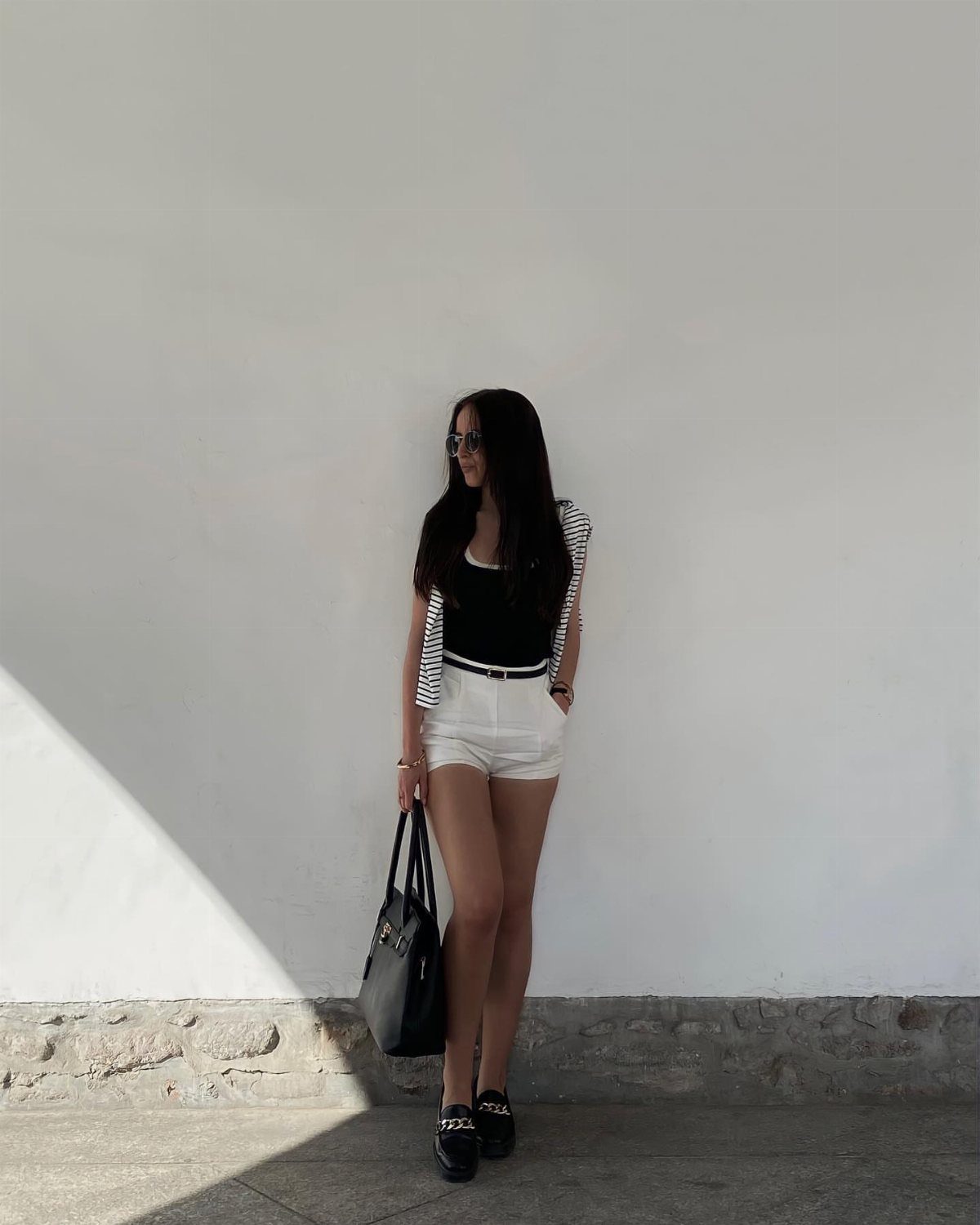 Woman in a black tank top and white shorts posing against a white wall.