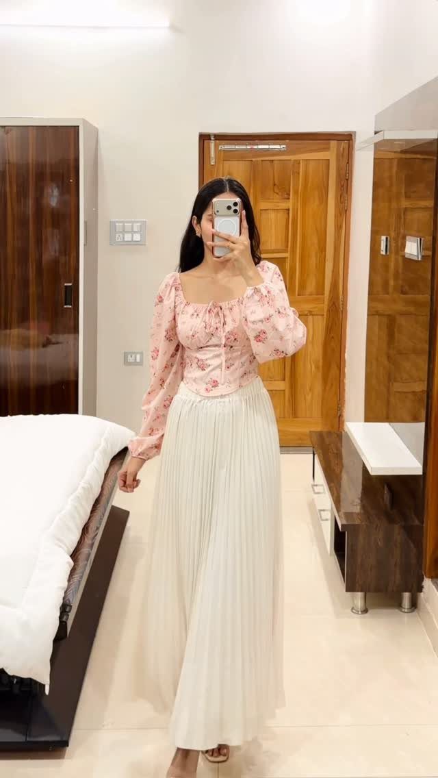 A woman posing in a mirror wearing a long white pleated skirt and floral blouse.