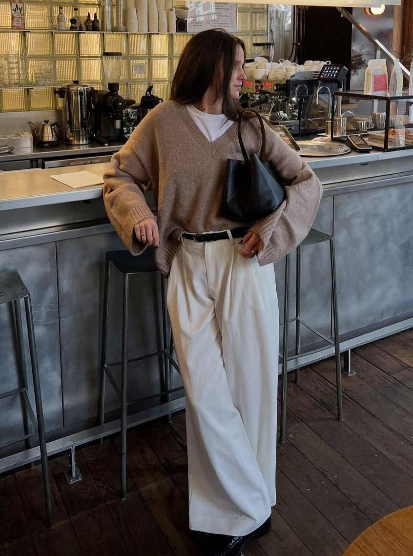 Woman in a tan heathered V-neck sweater and white wide-leg trousers standing in a cafe.