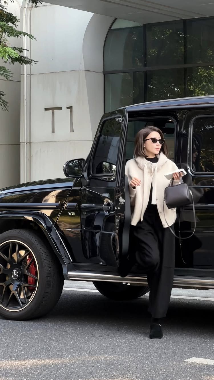 Woman in a cream textured sweater jacket and black trousers standing by a black SUV.