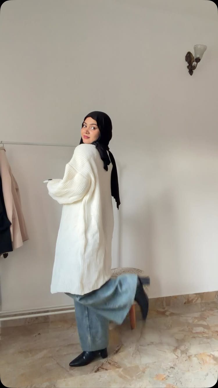 A woman in a long white tunic sweater and blue wide-leg pants posing indoors.