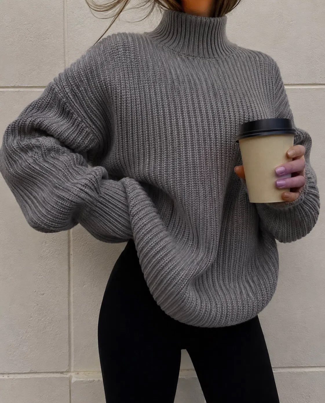 A person wearing a grey ribbed turtleneck sweater while holding a coffee cup.