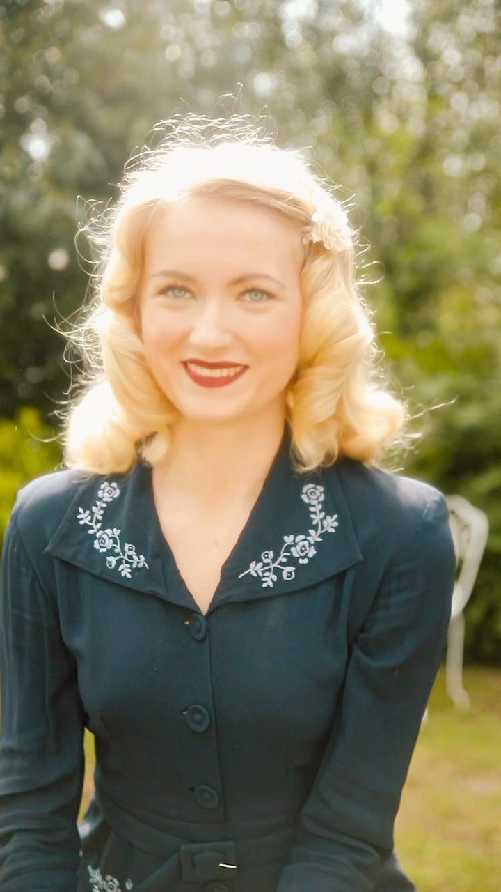 A blonde woman with vintage hair and red lipstick wearing an embroidered dark blouse.