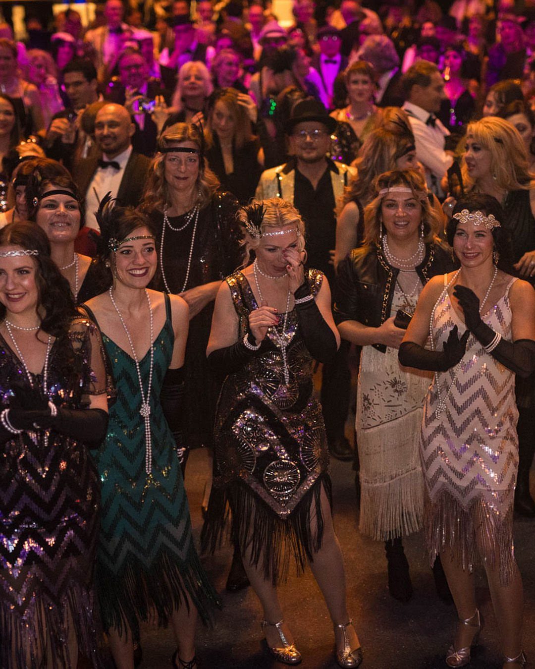 Women wearing sequined and fringed flapper dresses with pearl necklaces at a party.