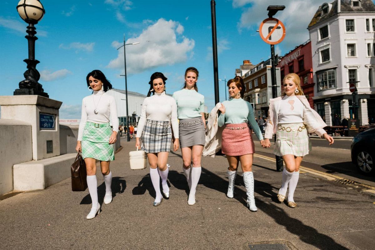Five women walking down a street wearing retro outfits and white knee-high boots.