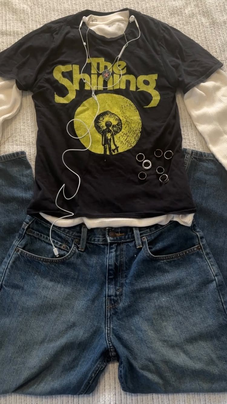 A black graphic t-shirt, white long-sleeve shirt, and blue jeans laid out on a grey surface.