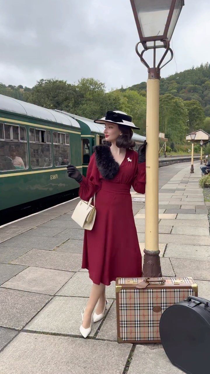 Woman wearing a red A-line dress and black hat standing on a train platform with a vintage suitcase.