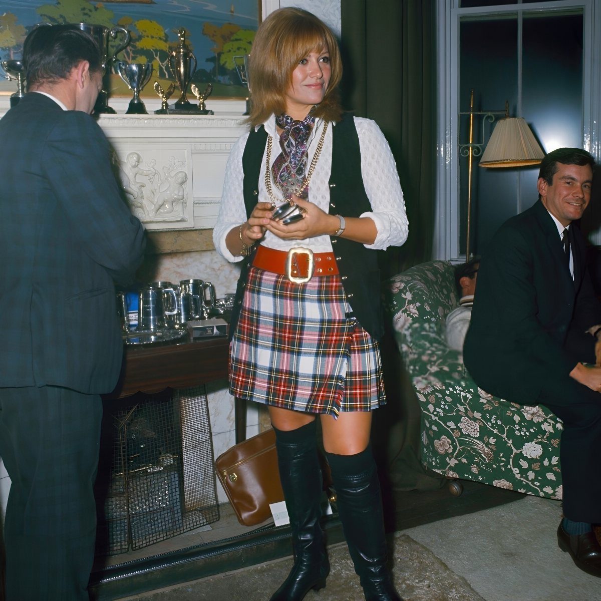 A woman wearing a plaid mini skirt, black vest, and tall black boots standing in a room.