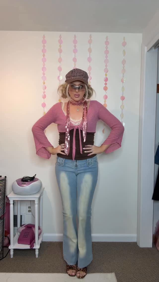 Woman posing in a pink flared top, corset, and patterned flared jeans with a newsboy cap.