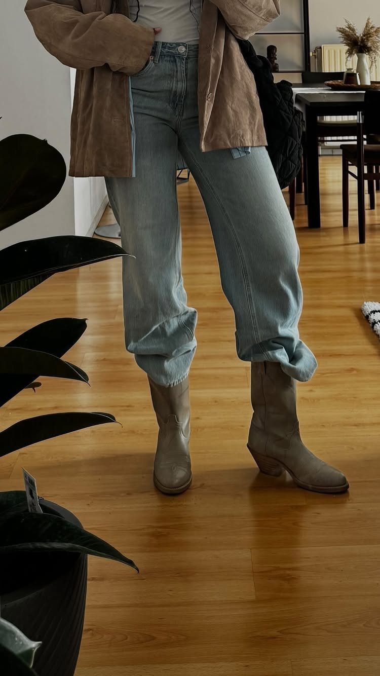 A person wearing light wash baggy denim jeans and tan boots standing on a wooden floor.