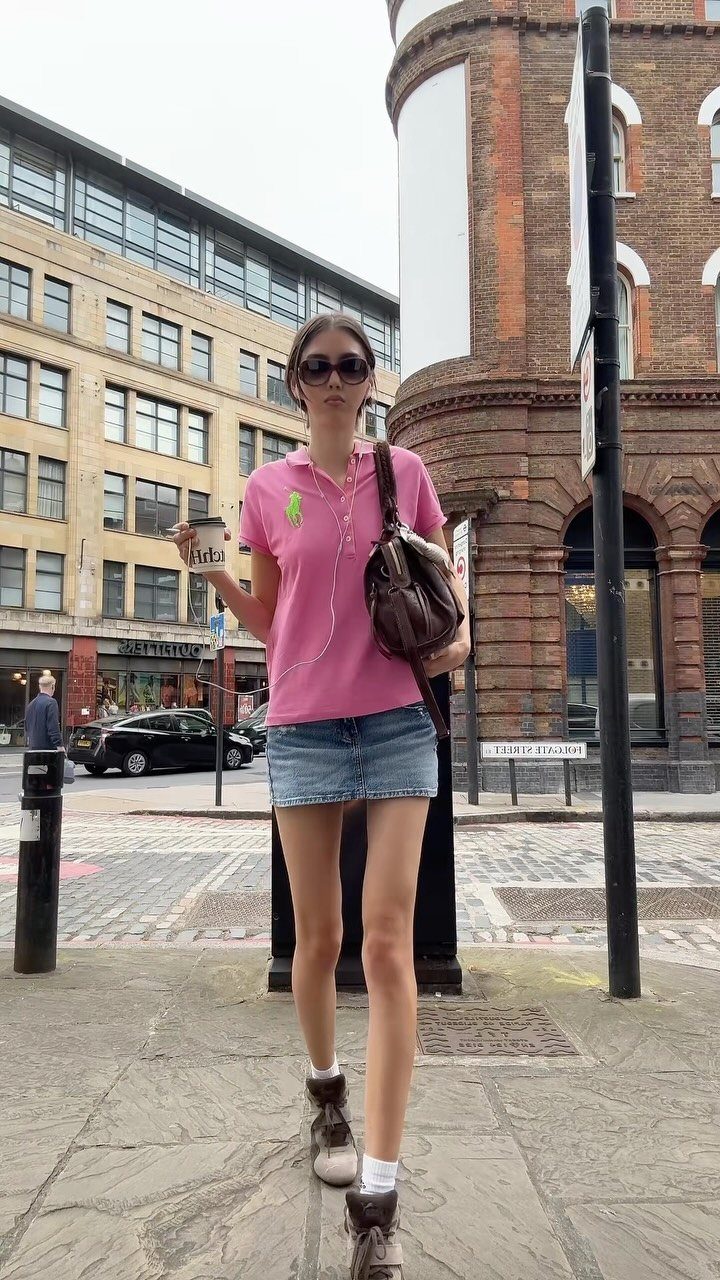 A woman in a pink polo shirt and denim mini skirt walking on a city street.
