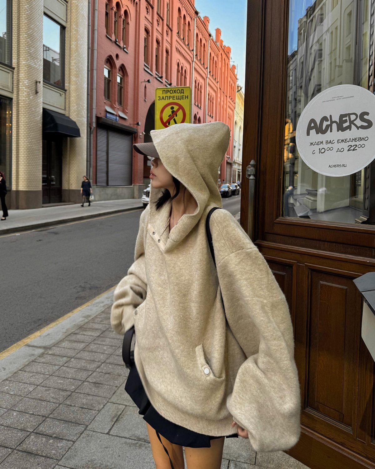 A person wearing a large beige oversized knit hoodie standing on a city sidewalk.