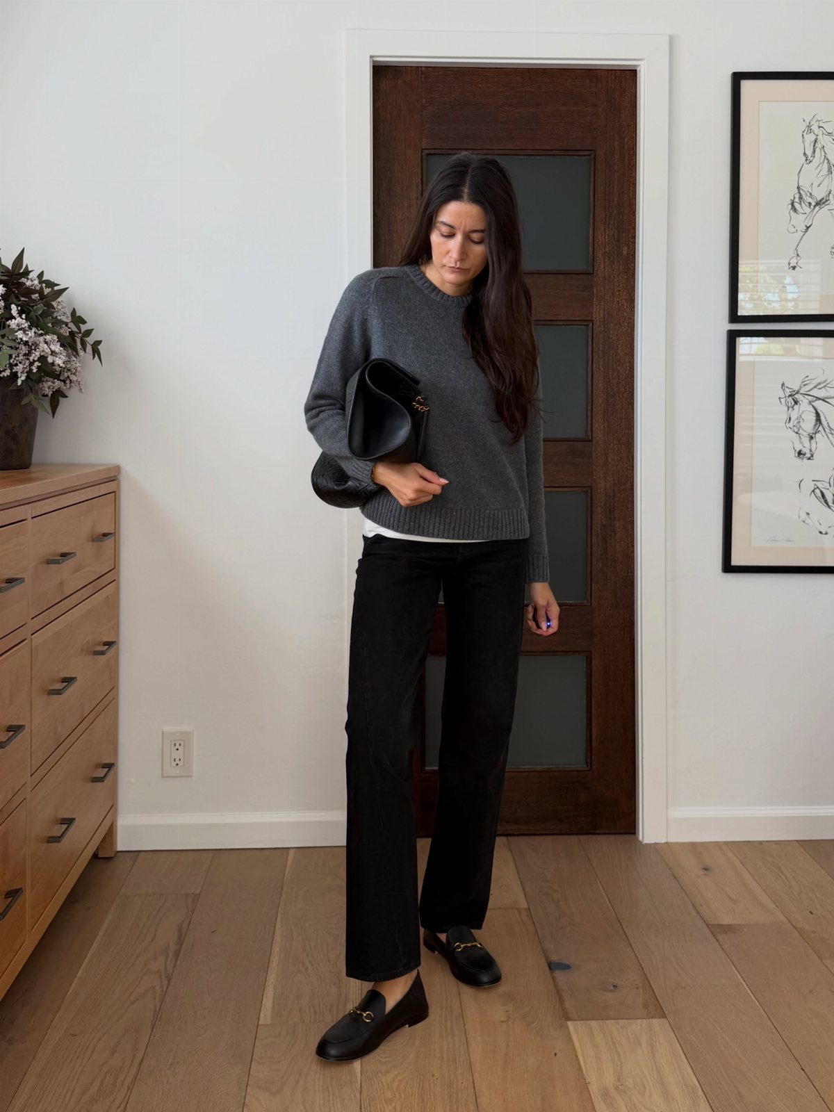 A woman wearing a grey sweater, black jeans, and black loafers standing indoors.