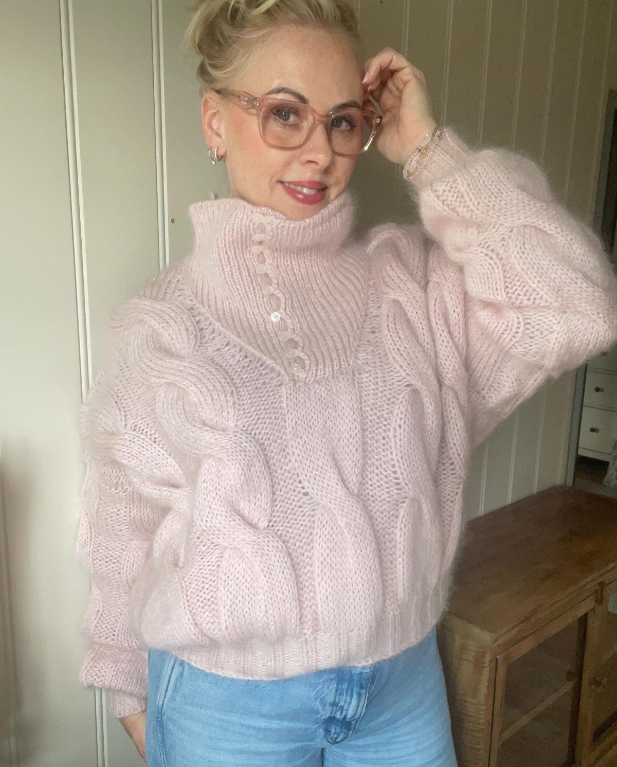 A woman wearing a thick, light pink cable-knit turtleneck sweater and blue jeans.