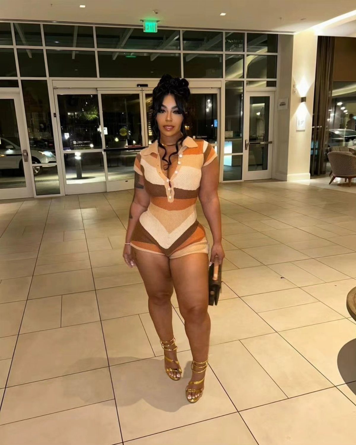 A woman posing in a patterned tan and orange romper and gold sandals.