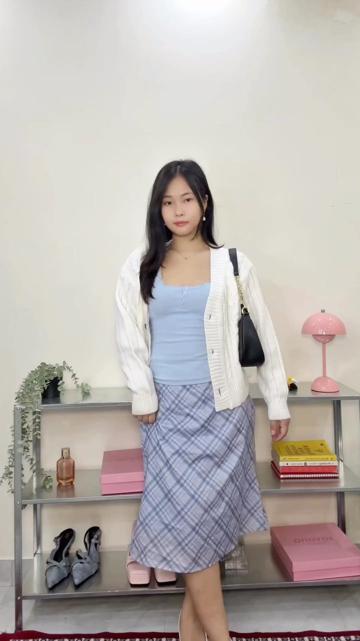 A young woman wearing a white cardigan, blue tank top, and plaid skirt posing indoors.