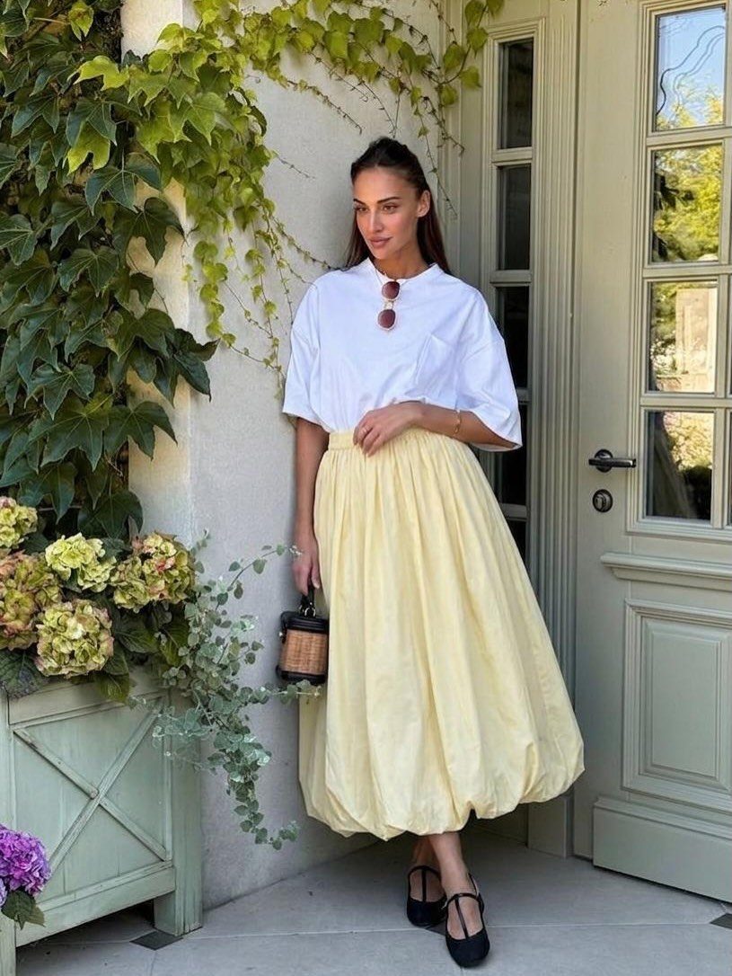 A woman posing in a white t-shirt and voluminous yellow skirt against a leafy green background.