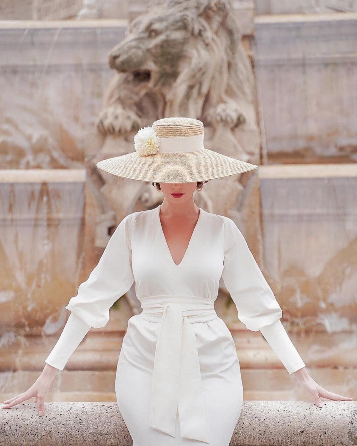 A woman in a white dress wearing a wide straw hat in front of a fountain.
