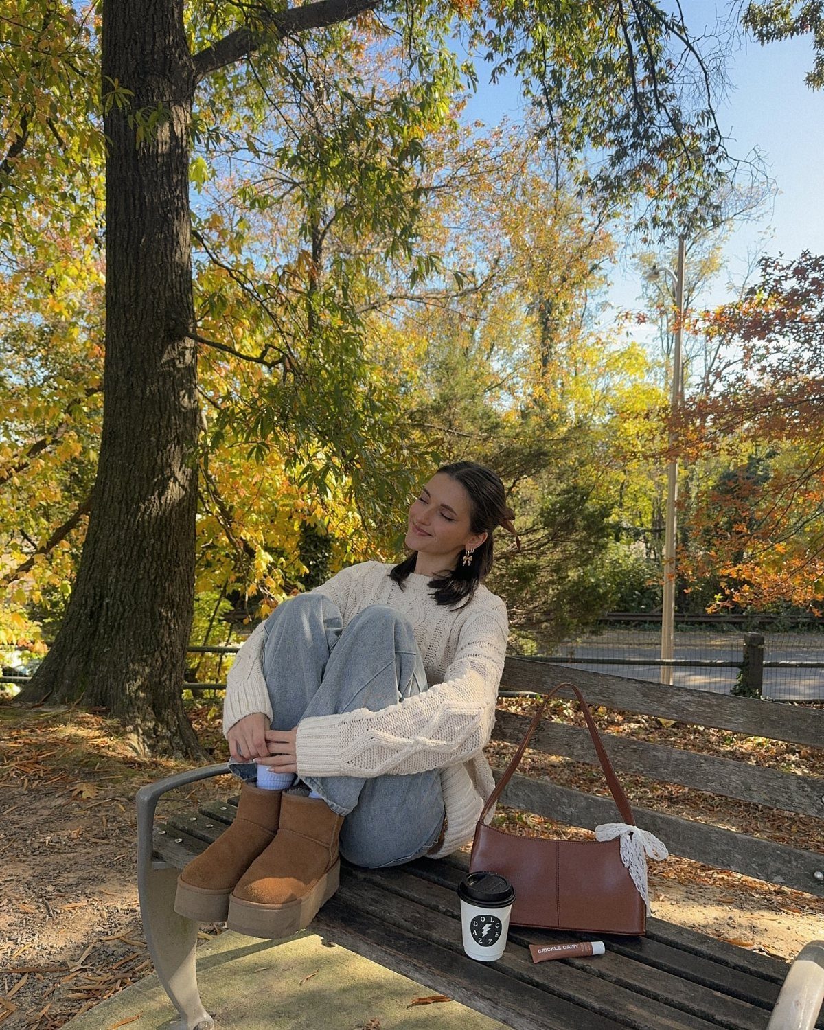 Woman in a white cable-knit sweater and wide-leg jeans sitting on a park bench.