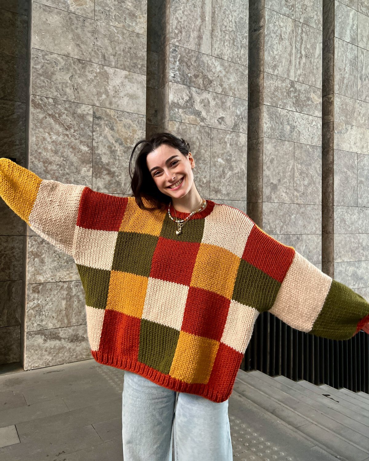A smiling woman wearing a colorful checkered knit sweater and light blue jeans outdoors.