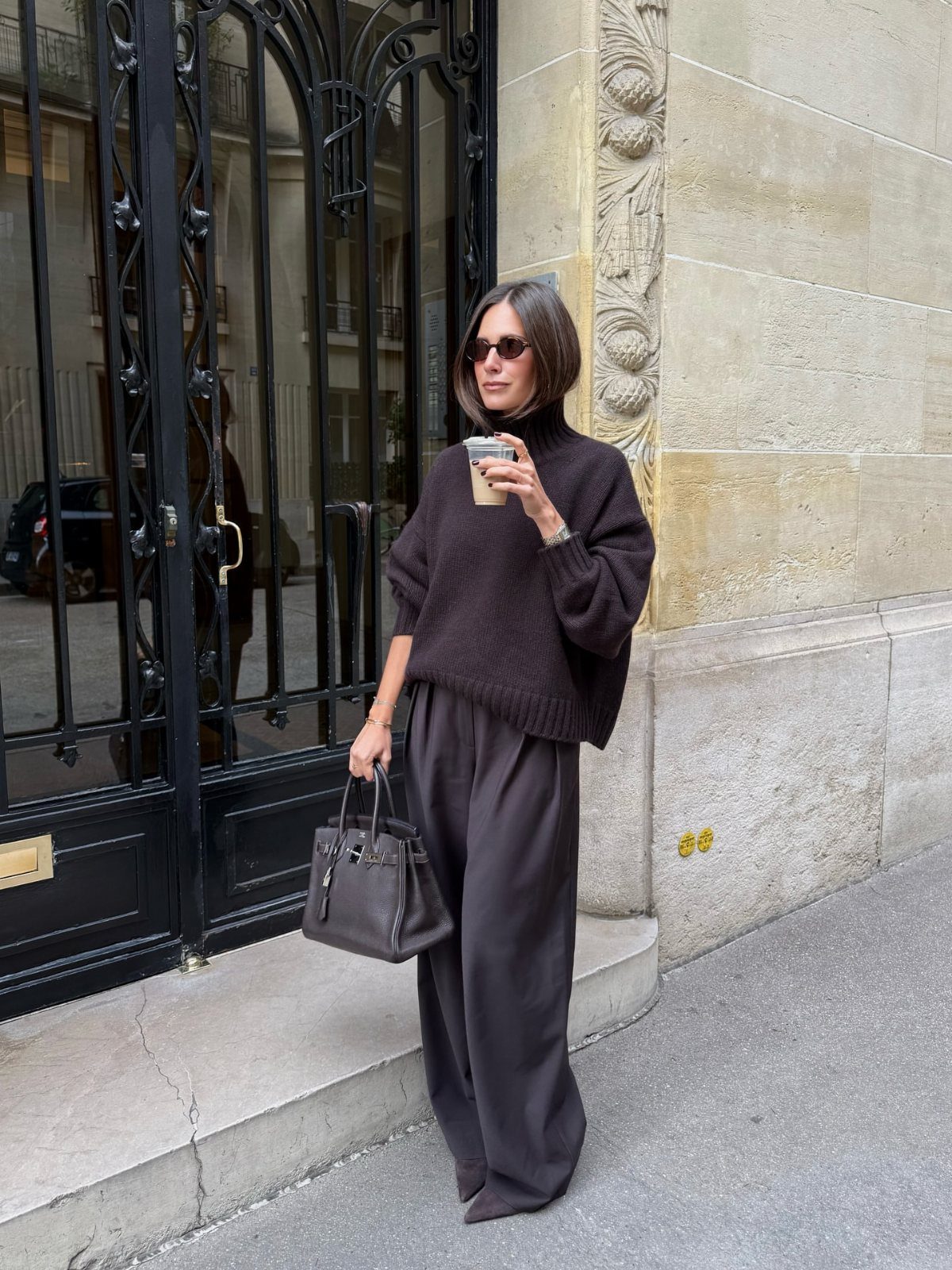 Woman in a dark brown chunky turtleneck sweater and wide-leg trousers standing outdoors.