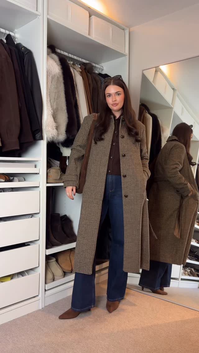 A woman posing in a closet wearing a long patterned coat and wide-leg blue jeans.
