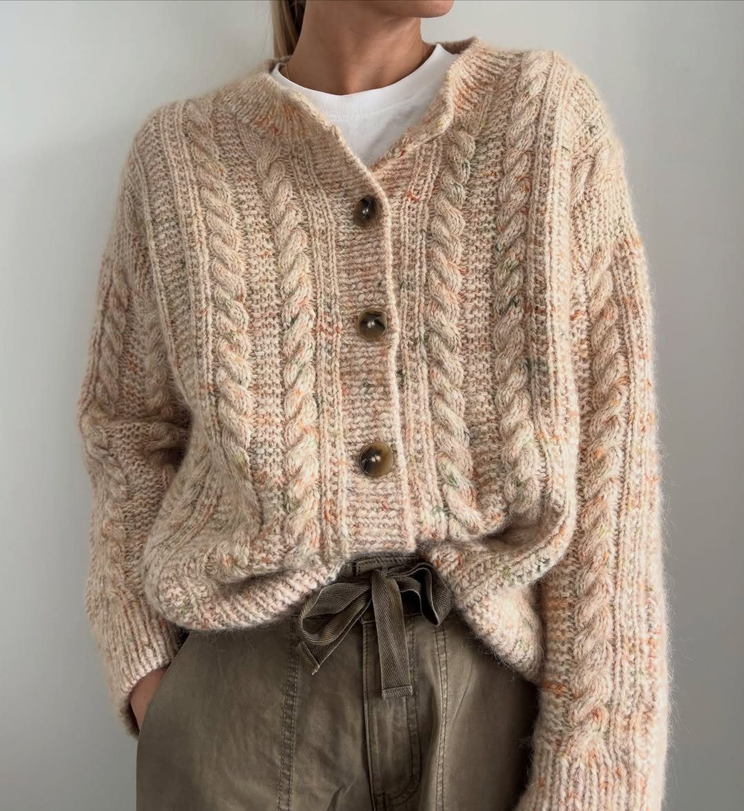 A person wearing a thick, cream cable-knit cardigan and olive green trousers.