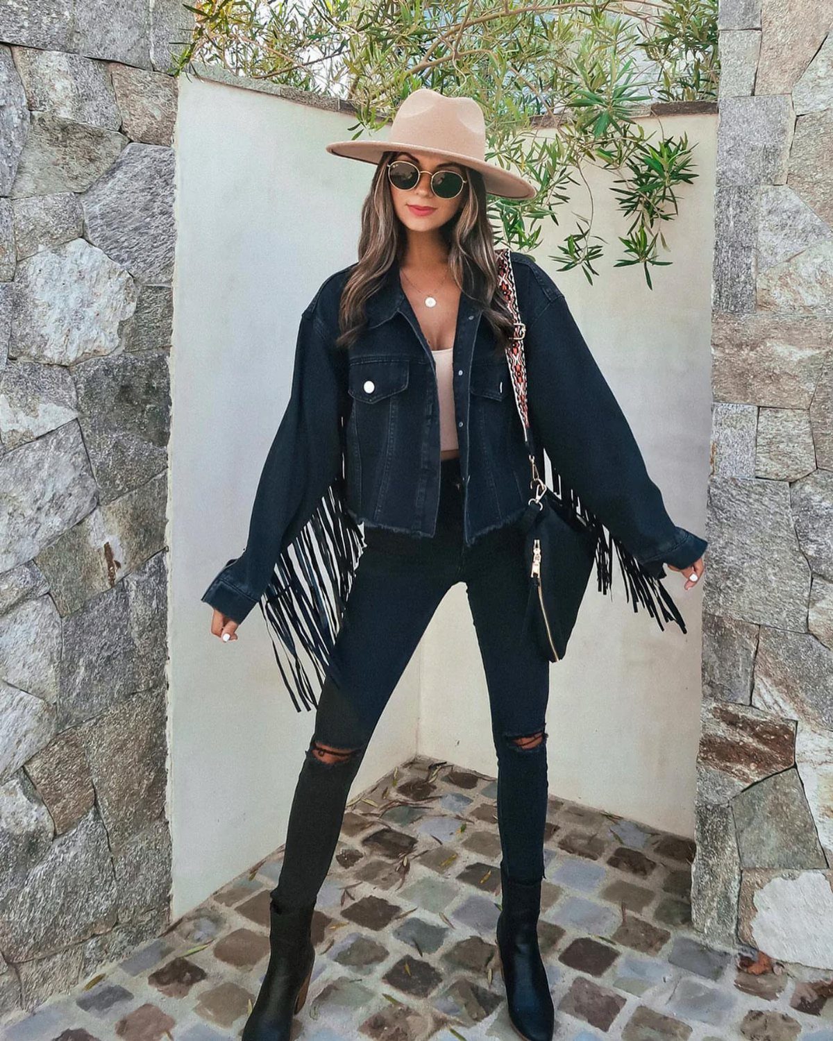 Woman wearing a black fringe denim jacket, black ripped jeans, and a tan wide-brimmed hat.