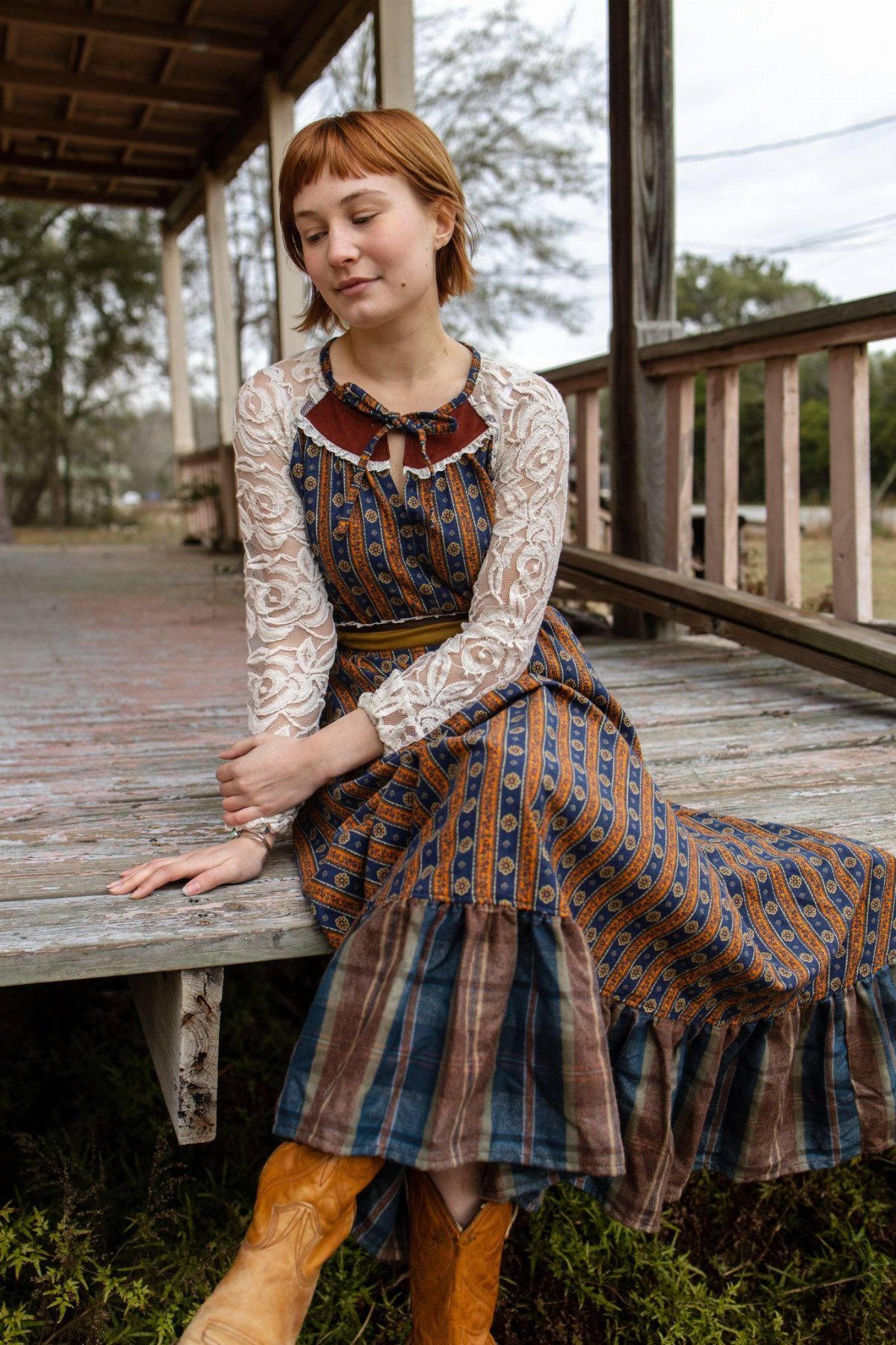 Woman in a long-sleeved patchwork prairie dress and cowboy boots sitting on a wooden porch.