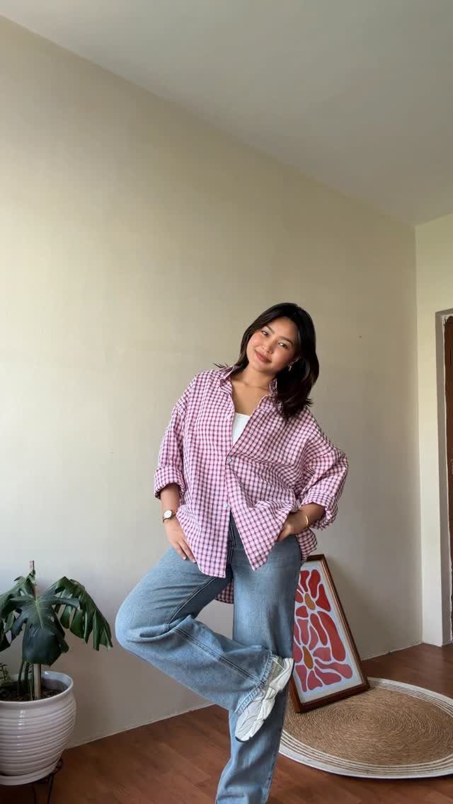 Woman wearing a pink checkered button-down shirt and light blue wide-leg jeans standing in a room.