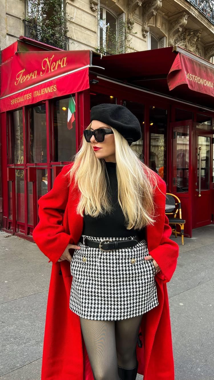 A blonde woman in a red coat and patterned skirt posing in front of a red building.