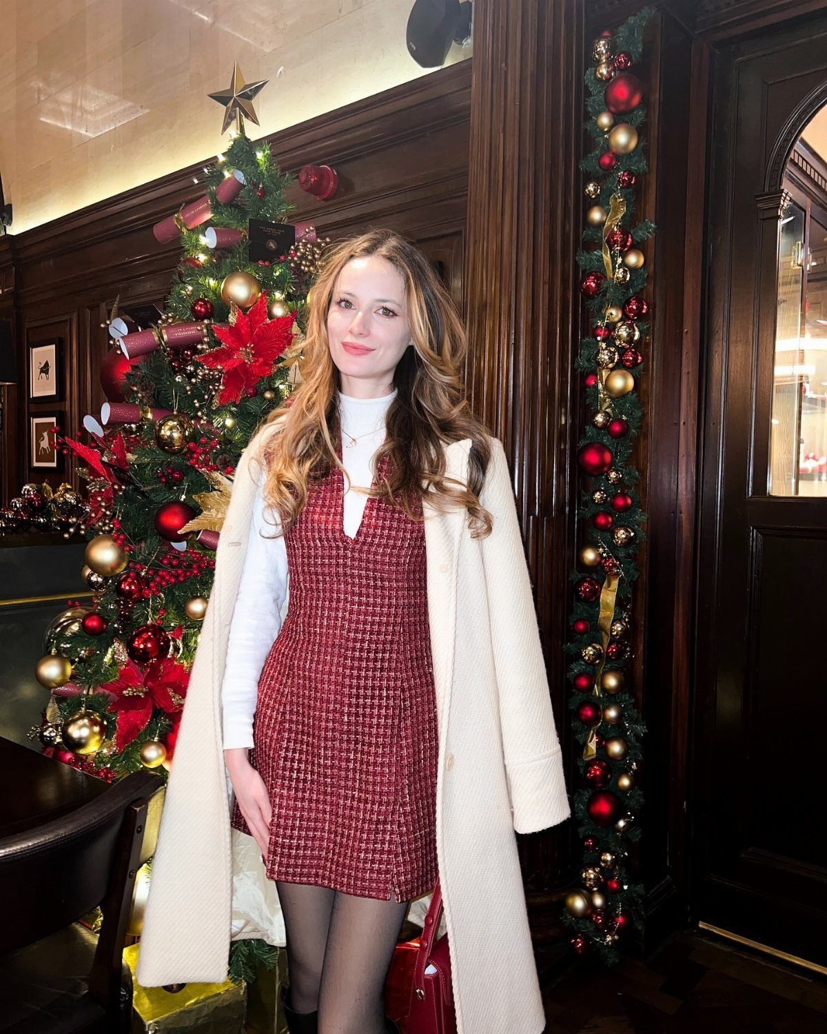 A woman wearing a red patterned dress and cream coat standing near a Christmas tree.