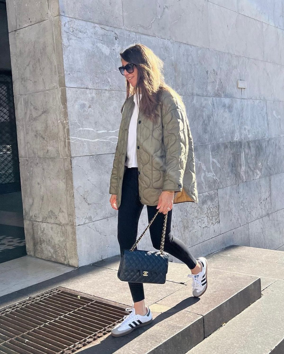 A woman walking in an olive green quilted jacket, black leggings, and white sneakers.