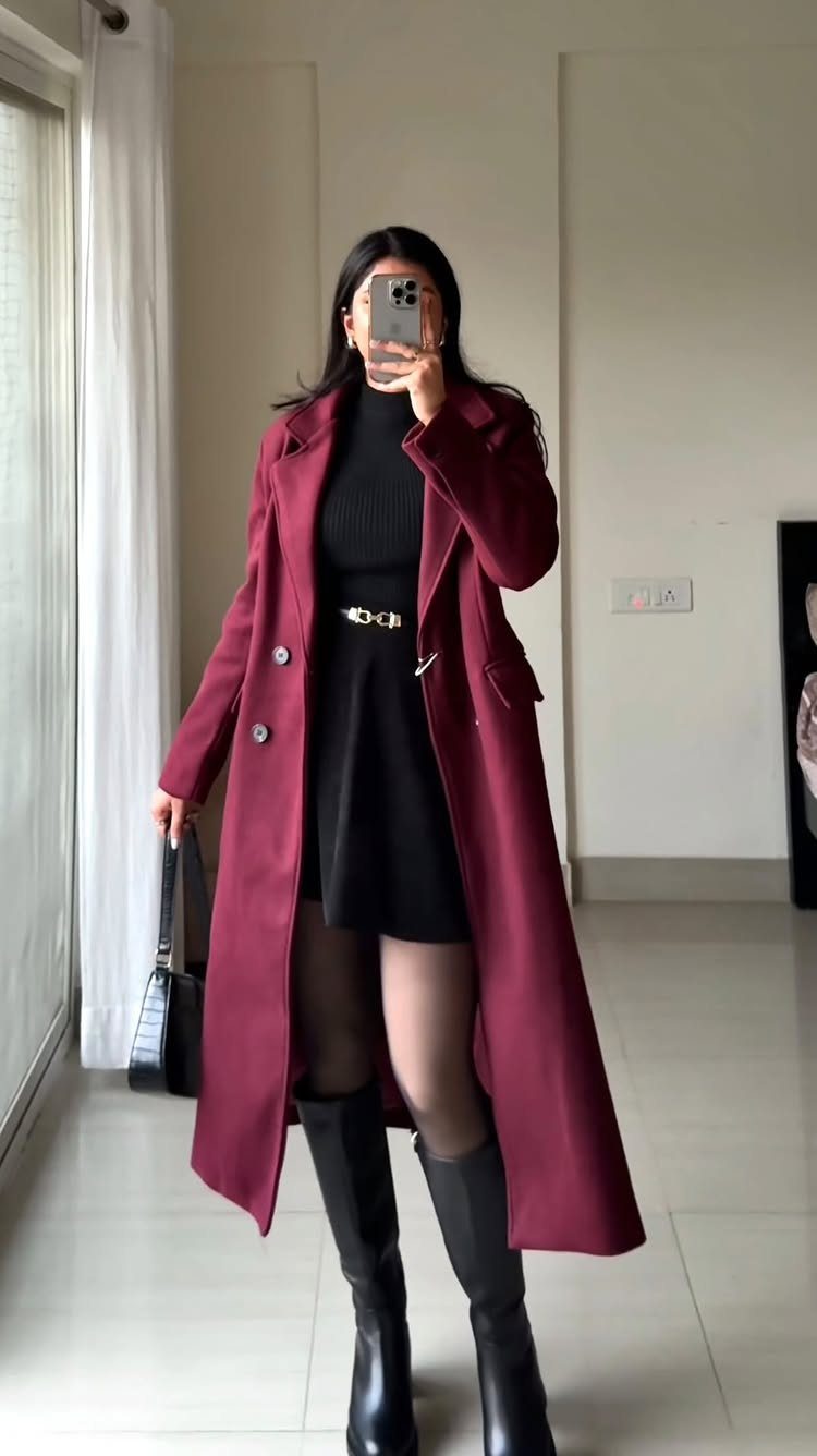 Woman in a burgundy coat and black sweater dress posing for a mirror selfie.