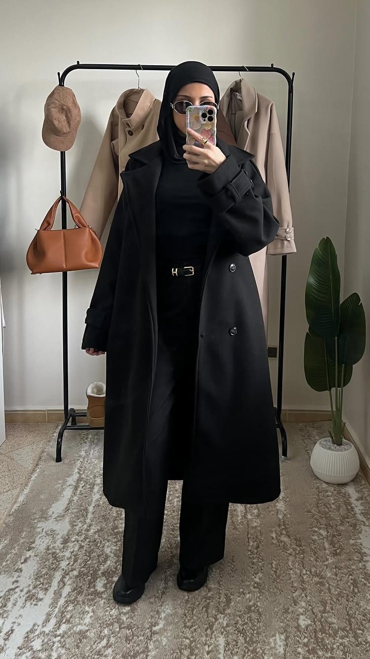 Woman in a black hijab and long black coat posing for a mirror selfie.