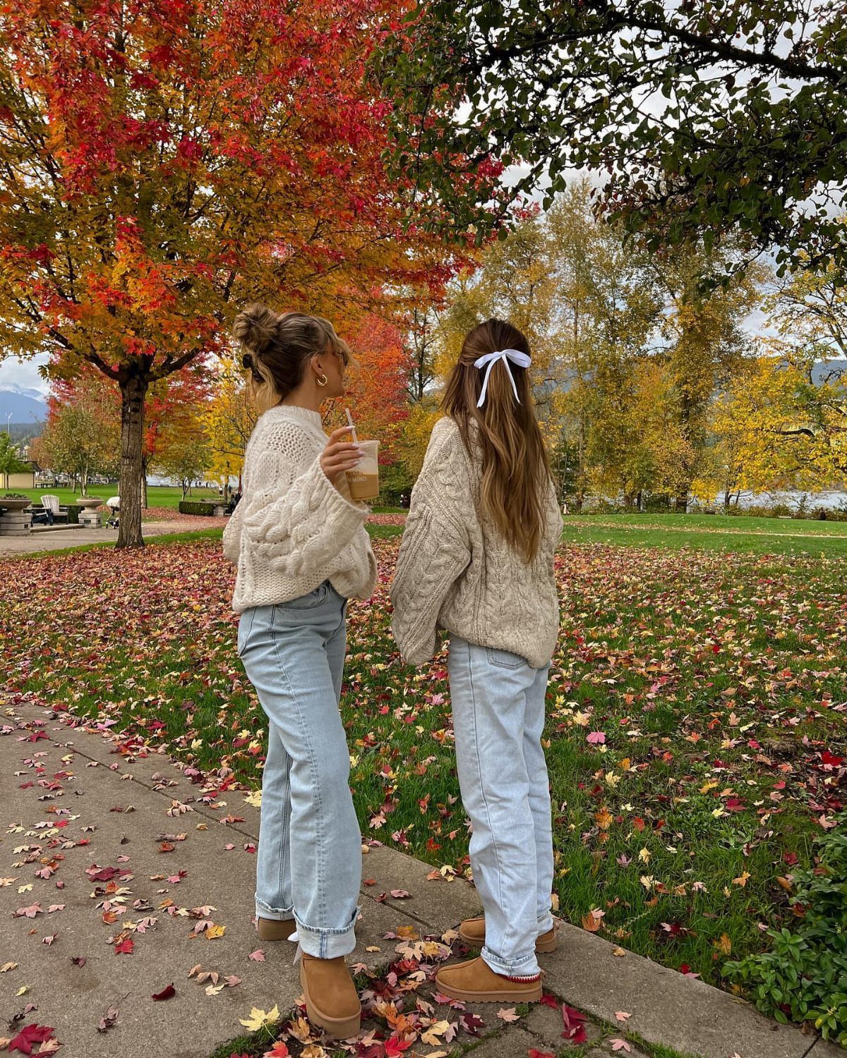 Two women wearing chunky cable-knit sweaters and light blue jeans standing in an autumn park.