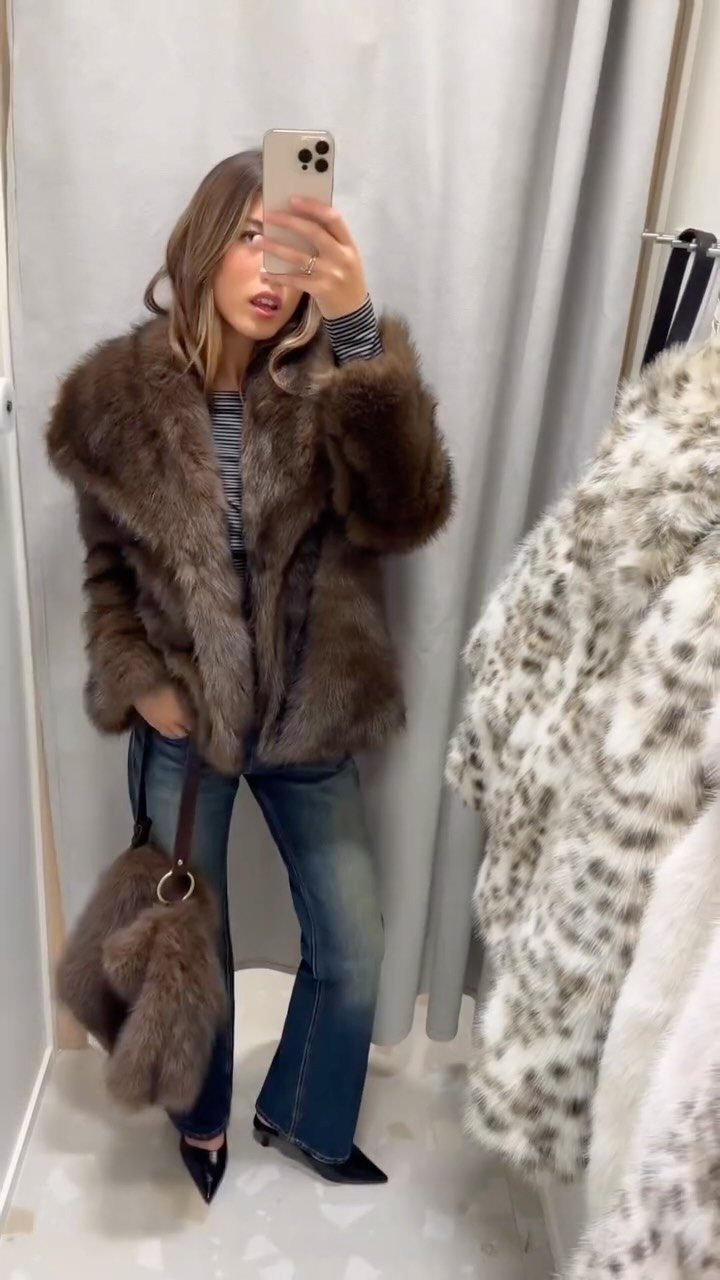 Woman in a brown fur coat, striped shirt, and blue jeans taking a mirror selfie.