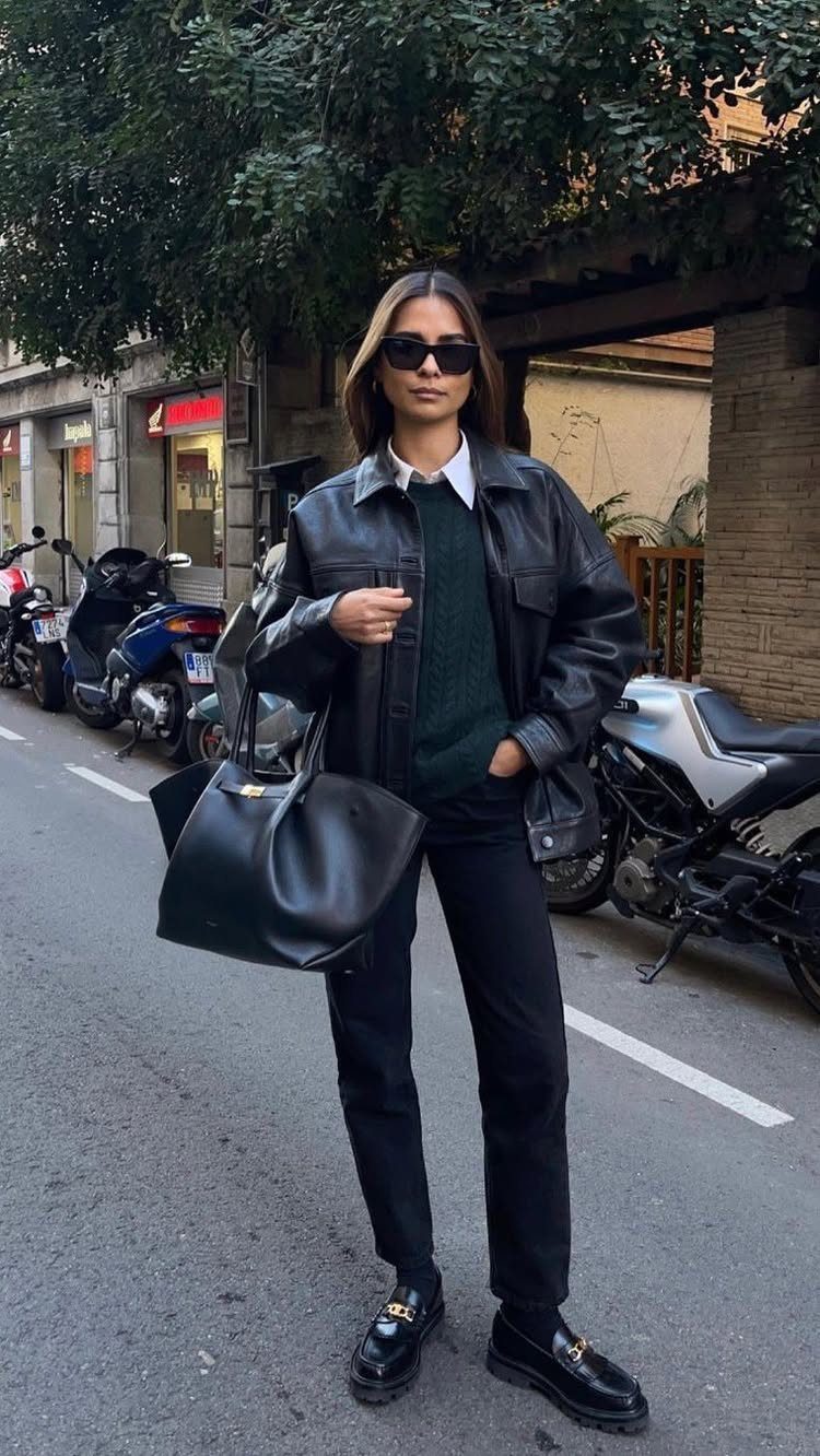 A woman in a black leather jacket, green sweater, and black trousers standing on a city street.