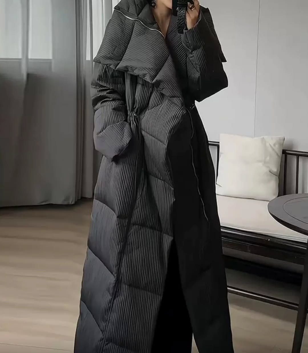 A person wearing a long, black quilted oversized puffer coat indoors.