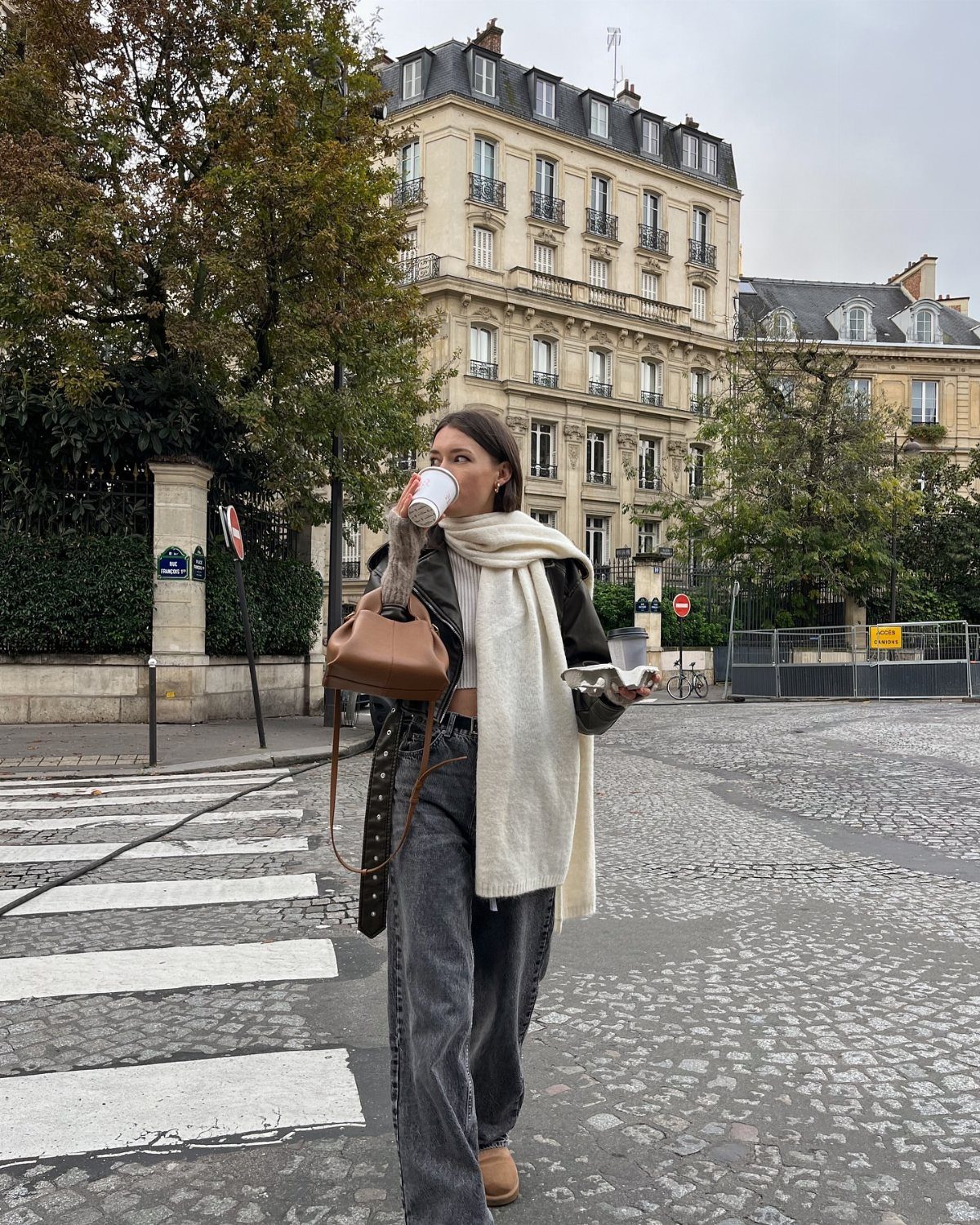 A woman in a cream scarf and dark jeans walks across a city street holding a coffee cup.