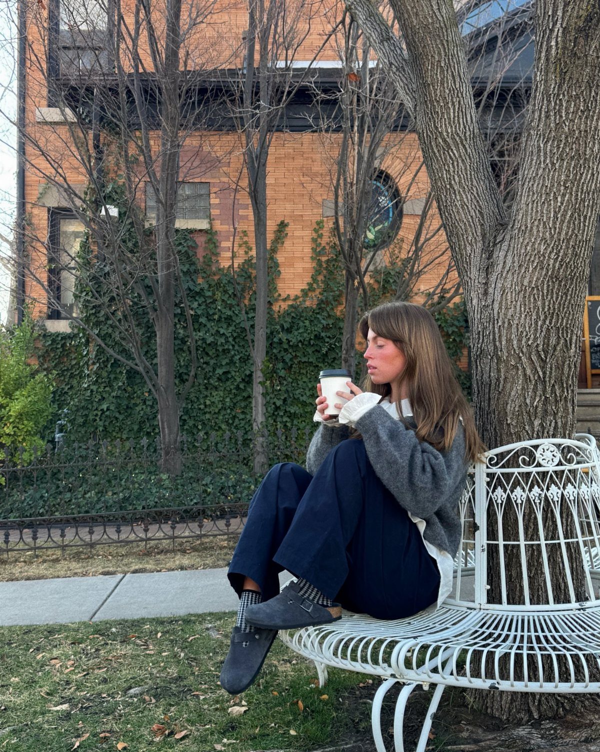 Woman in a grey sweater and dark wide-leg trousers sitting on a white metal bench.