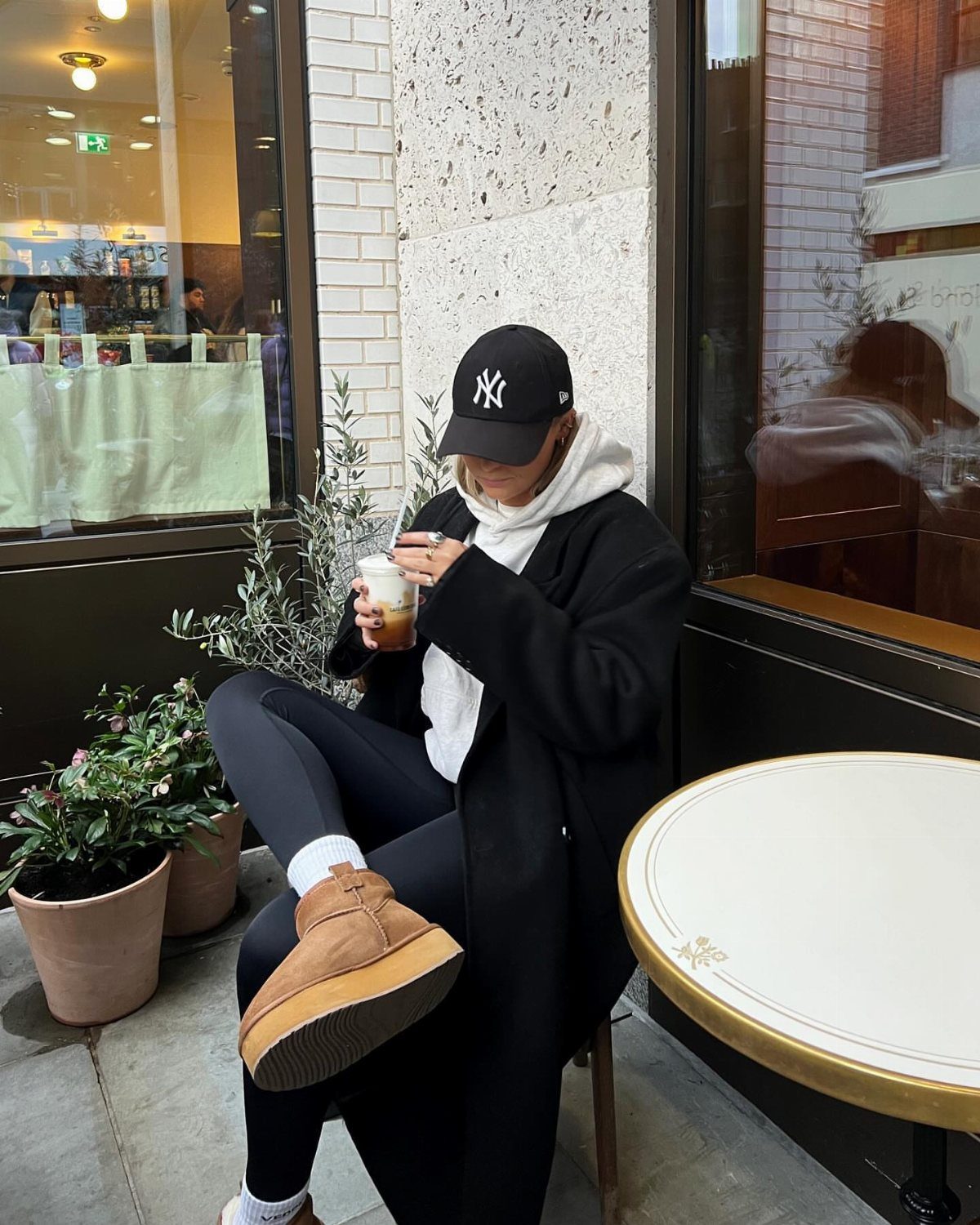 A person in a hoodie, black coat, and leggings sitting at an outdoor cafe table.